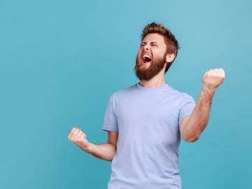 portrait of overjoyed bearded man standing with excited expression raising fists screaming shouting yeah celebrating his victory success