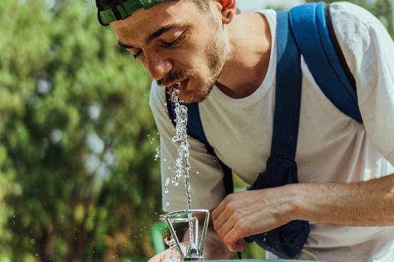 a shallow focus of a young man wearing a cap and drinking water from a drinking fountain in a park