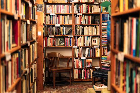 an array of book titles under a variety of category headings on bookshelves above a wooden chair in the corner of a san francisco second-hand bookstore