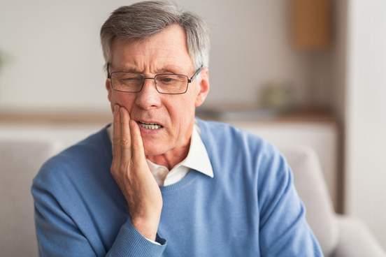 elderly man having toothache touching cheek suffering from pain sitting on sofa at home