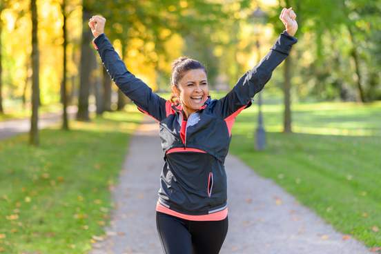 happy fit middle aged woman cheering and celebrating as she walks along a rural lane through a leafy green park after working out jogging