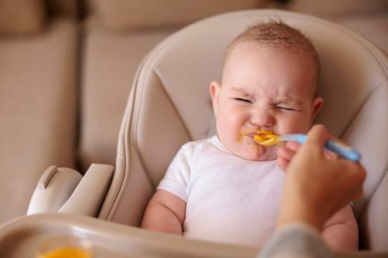 little baby boy sitting in high chair making funny faces and refusing to eat pumpkin porridge while mother trying to feed him using spoon