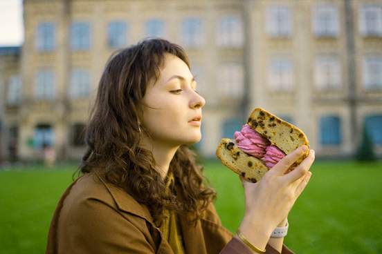 young caucasian woman sitting on lawn and eating huge sandwich made of biscuit and marshmallow