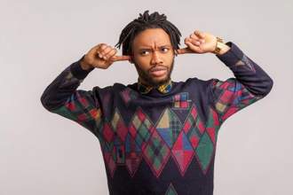 a man in a patterned sweater childishly placing his pointer fingers in his ears while looking upset