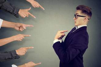 a man looking shocked pointing at himself as a group of hands on the left side of the screen are all pointing directly at him