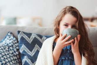 a young girl sitting on a couch wrapped in a blanket while sipping a drink from a large mug and staring straight at the audience