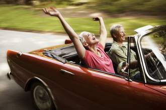 an older couple driving in a convertible with the top down and the woman in the passenger seat is enjoying herself with her hands lifted in the air