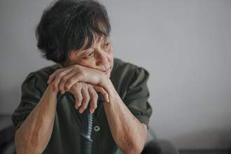 an older lady leaning forward with her hands propped up on her cane as she looks sadly to the right of the screen