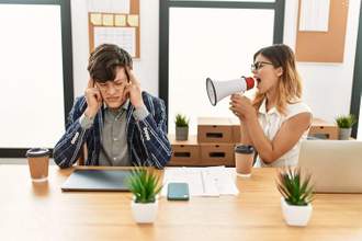 at the office two workers are sitting next to each other the female is yelling into a megaphone while the male is holding his head looking upset