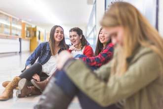 girl sitting alone in the hallway while a group point and laugh at her