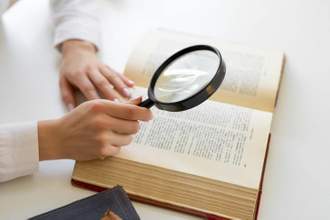 hands holding a magnifying glass looking at an open book