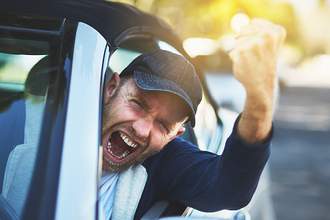 man angrily shaking fist behind wheel of a car