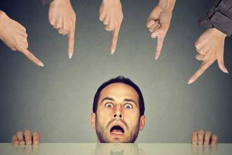 man ducking behind desk looking shocked while fingers are pointing directly at him from above