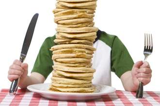 man holding a large fork and knife while a huge stack of pancakes sit in front of their face