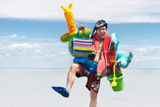 man in a bathing suit holding all sorts of beach accessories walking down the beach