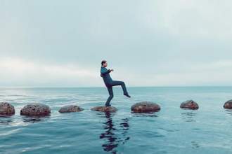 man in a suit tiptoeing on large rocks across a vast body of water