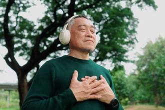 man peacefully meditating with his eyes closed and arms to his chest as he stands under a tree listening to music