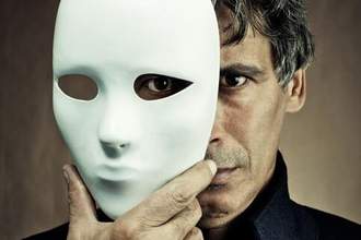 menacing looking man holding a white mask partially in front of his face