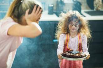 mother cwith her hands on her head looking distraught while her daughter is holding a smoking black baked good