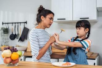 mother holding a fork with food on it to unwilling daughter