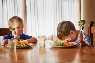 two children with plates full of vegetables looking miserable while trying to eat broccoli