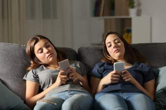 two girls sitting on a couch staring at their phones looking bored