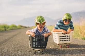 two kids with watermelons on their heads rolling down a hill on a skateboard