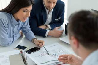 woman leaning over a table pointing out and explaining paperwork to two coworkers