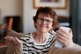 woman looking directly at the audience holding her phone in one hand and shrugging her shoulders