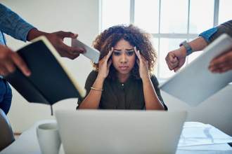woman sitting in front of her computer looking concerned with multiple hands reaching out from the sides of the screen trying to hand her more work