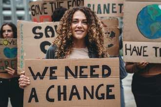 woman standing in the middle of the frame smiling at the audience while holding a sign for change in regards to helping the environment