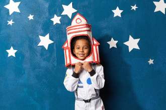 young child dressed an astronaut with his head inside a small paper rocket standing in front of a star filled sky background