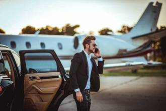 young rich businessman talking on a mobile phone while getting out of a luxurious car parked next to a private airplane on a tarmac