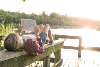 young teen laying down reading a book by the water